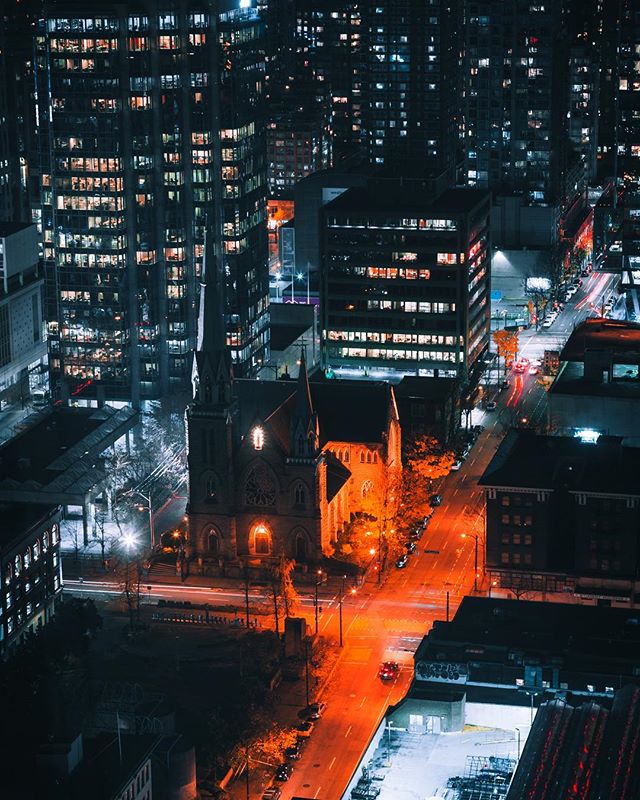 Night view of a city featuring illuminated buildings, a church, and a street with light trails.