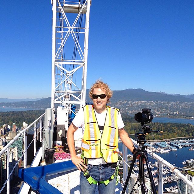 A construction worker stands with a camera on a crane overlooking a city.