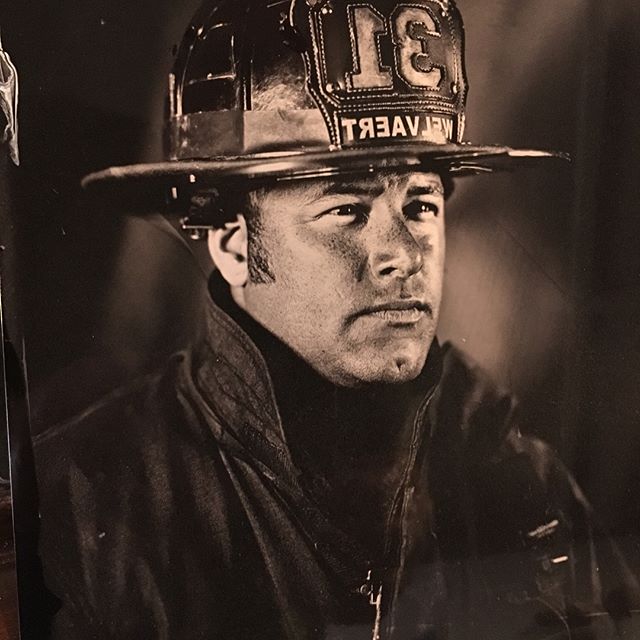 A black and white portrait of a firefighter in uniform and helmet, looking serious.