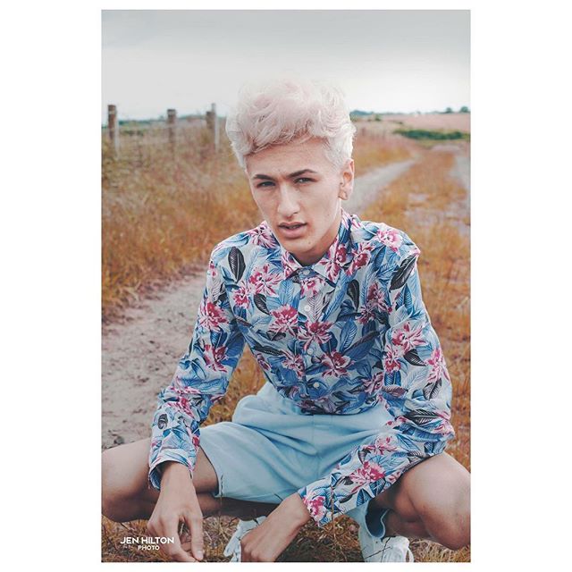 A young man with light hair poses in a floral shirt and shorts in a rural field.