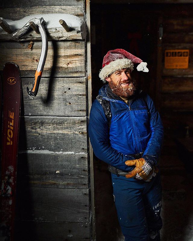 Man wearing a Santa hat stands next to skis and an ice axe in a cozy cabin setting during the winter.