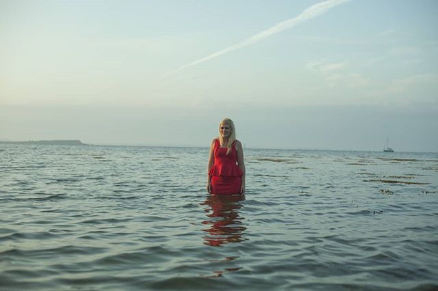 A woman in a red dress stands serenely in the ocean water under a bright sky.