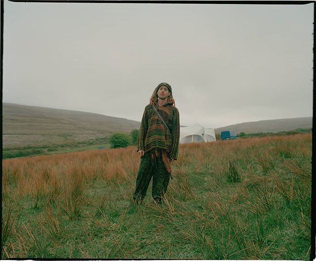 A man stands in a grassy field, wearing a poncho, with hills in the background.