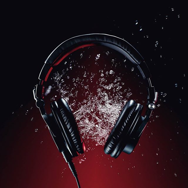 A set of black headphones suspended mid-air, surrounded by water droplets, set against a dark red gradient backdrop.