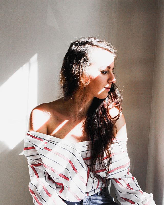 A brunette woman poses indoors in soft light wearing an off-the-shoulder striped shirt.