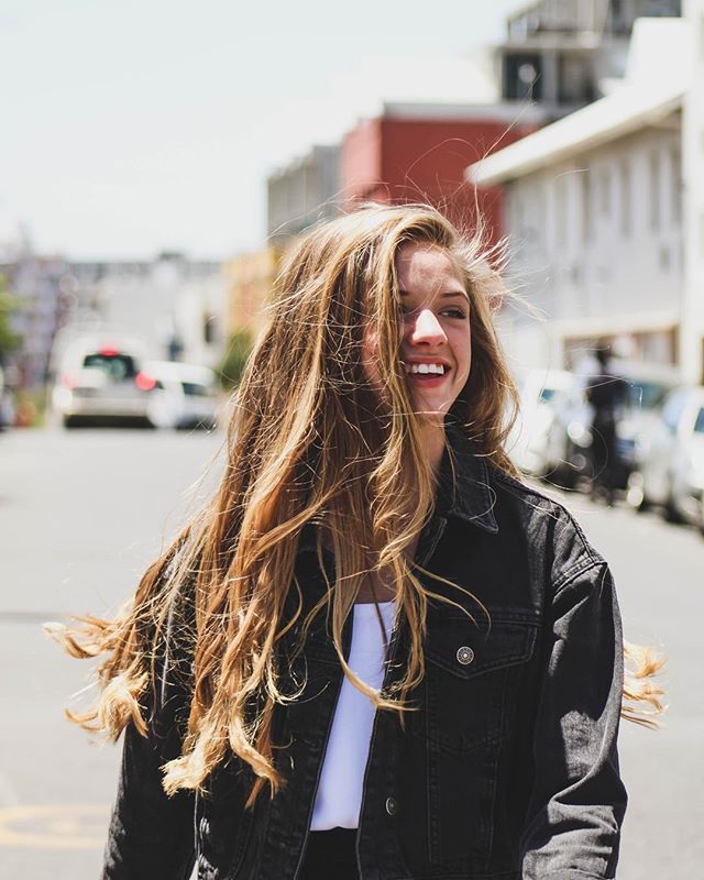 A smiling woman with blonde hair walks on a city street wearing a jacket.