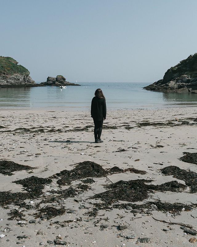A lone woman stands on a sandy beach, gazing out at the calm ocean, islands visible in the distance, creating a tranquil scene.
