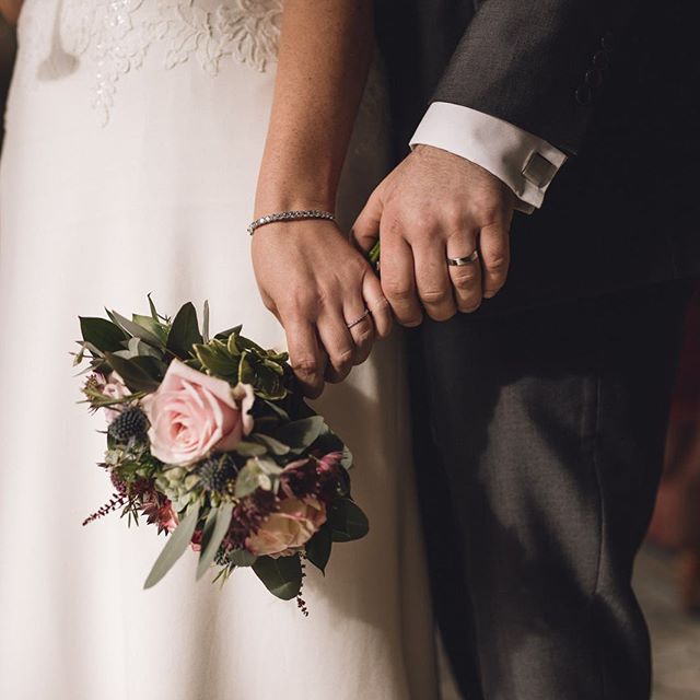 A bride and groom hold hands displaying their wedding rings and a lush bouquet.