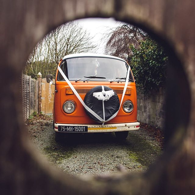 An orange VW bus, decorated for a wedding, is seen through a tunnel. The background is a rural landscape.