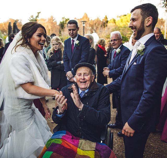 A bride and groom share a joyful moment with wedding guests and a man in a wheelchair during an outdoor wedding celebration.