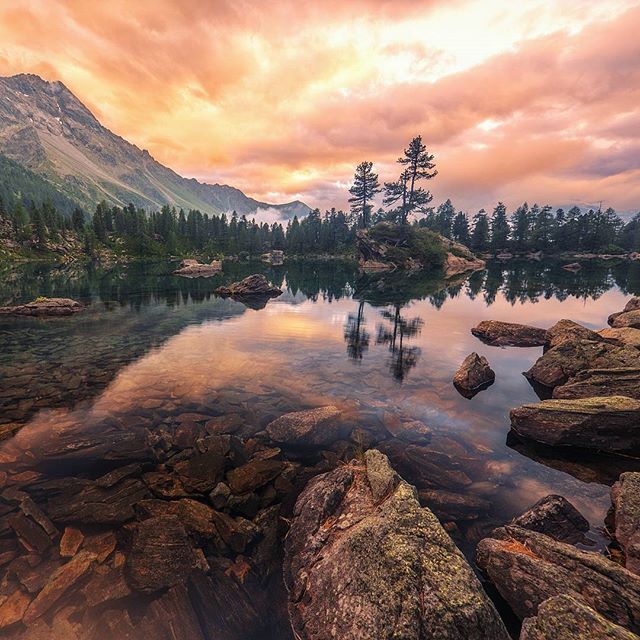 A scenic view of a tranquil lake reflecting the sky and surrounding mountains and forest, with rocks in the foreground.