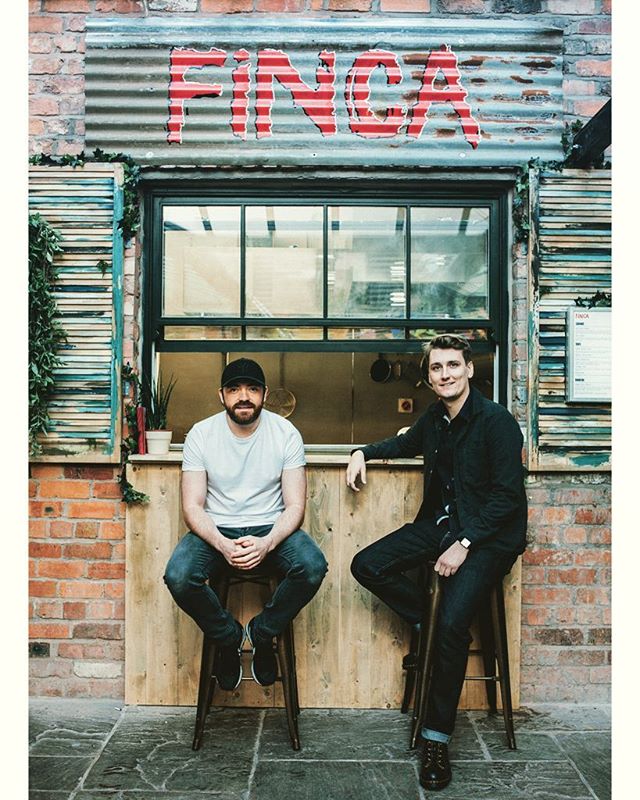 Two men are sitting on stools in front of Finca restaurant, posing for a photo.