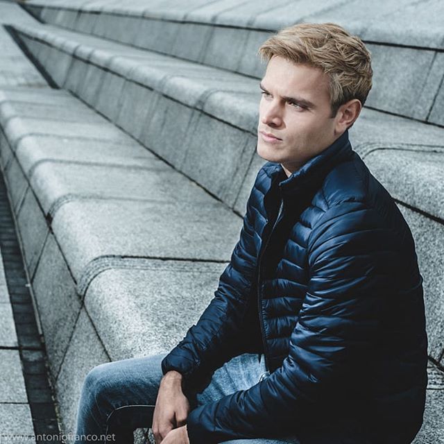 A man with blonde hair sits on concrete stairs wearing a blue jacket and jeans.