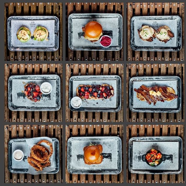 A variety of gourmet dishes including burgers, avocado toast and onion rings artfully arranged on trays over a rustic wooden background.