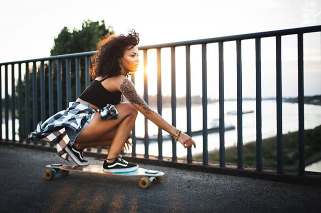A young woman with curly hair rides a skateboard on a bridge with a river view during a sunny day.