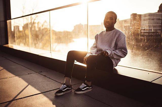A young man sits on a rooftop ledge in the city, wearing sneakers and a sweatshirt.