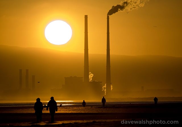 People walk on a beach silhouetted by smokestacks and a setting sun, emphasizing environmental issues and industrial impact.