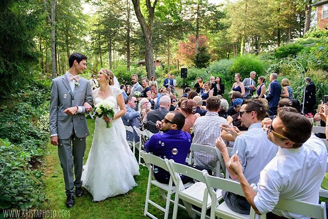 A newly married couple walks down the aisle as guests celebrate with bubbles at an outdoor garden wedding.
