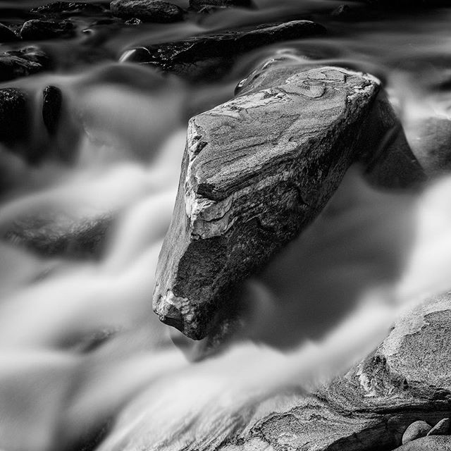 A black and white long exposure captures a rock surrounded by flowing water in a peaceful outdoor scene.