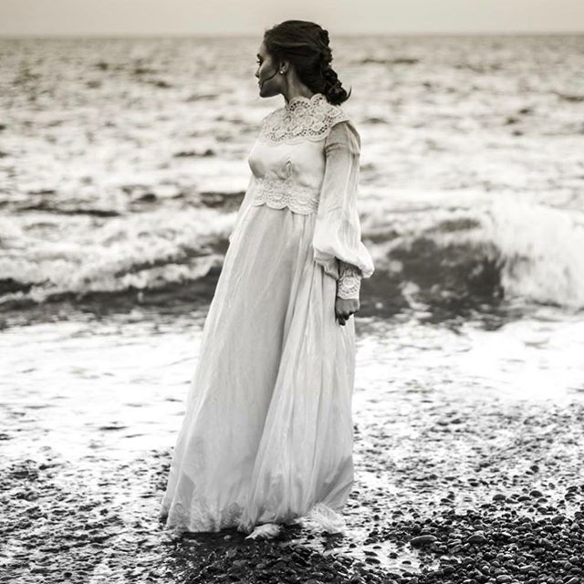 A woman in a vintage dress stands on a rocky beach by the ocean, in an elegant and timeless scene.