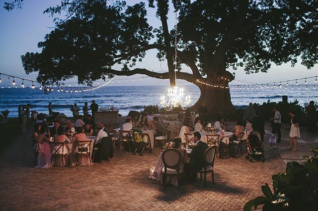 A romantic wedding reception unfolds by the ocean under a string of lights and a chandelier.