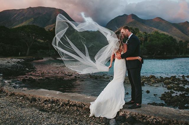 A bride and groom share a kiss on a beach with mountains in the background, the veil blowing in the wind.