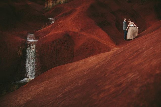 A couple stands together on a red desert landscape near a waterfall, creating a romantic and intimate moment.