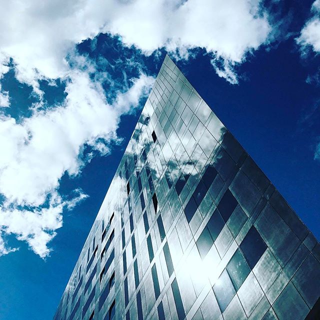 A modern glass building reaches towards a bright blue sky with fluffy white clouds, reflecting the sky on its surface.