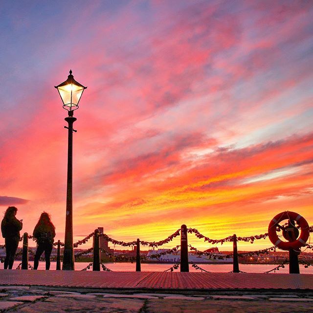 People watch a vibrant sunset over the waterfront, framed by a lamp post and lifebuoy on a scenic dock.