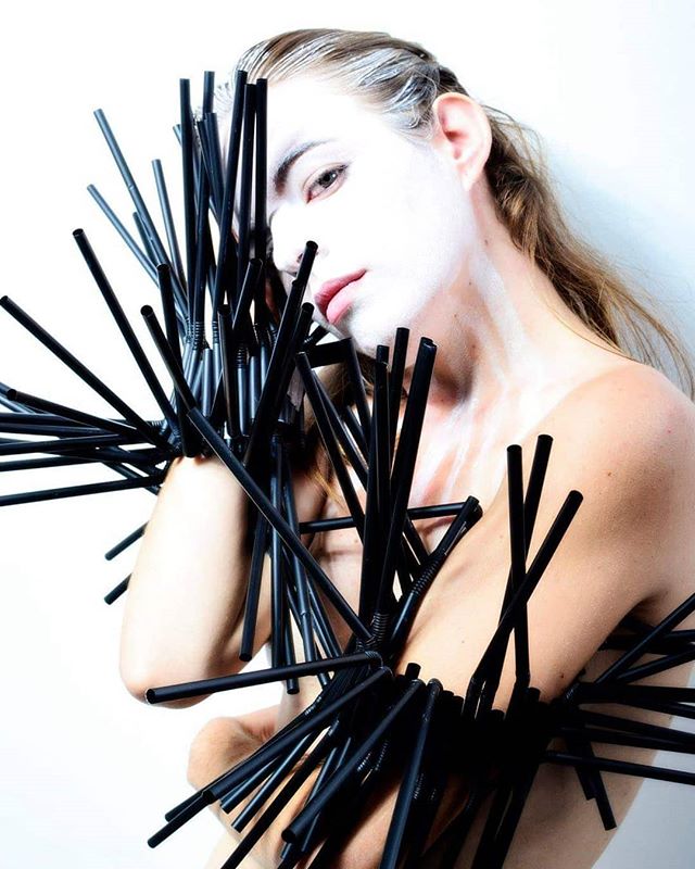 A woman with white makeup poses wearing an arrangement of black straws in an avant-garde fashion concept.