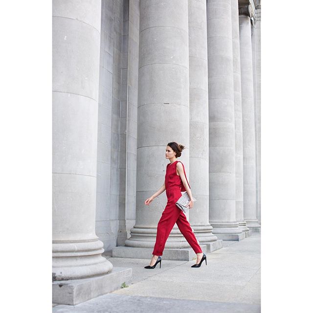 A woman in a red jumpsuit walks confidently past a row of grand columns.