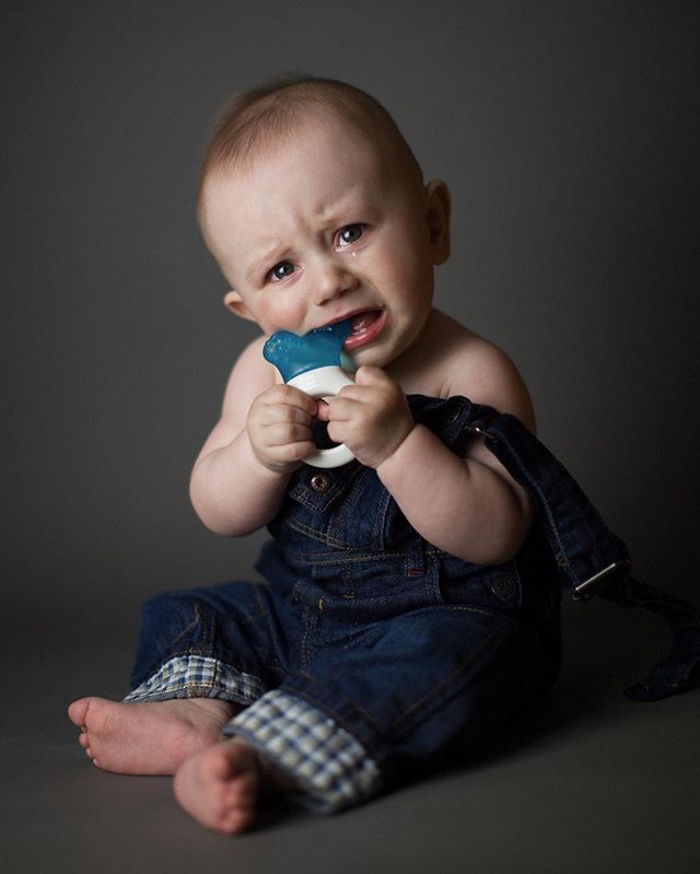 A crying baby boy in denim overalls holds a teething toy in a studio shot against a gray background.