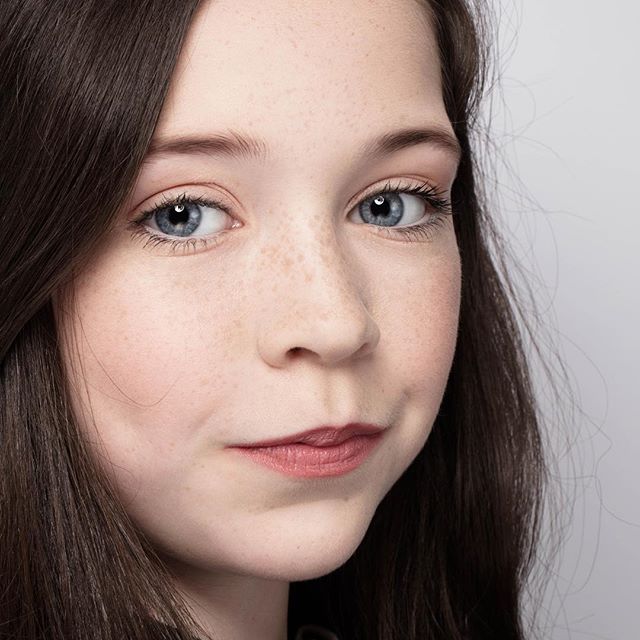 A close-up portrait of a young girl with blue eyes and freckles, posing in a studio setting.