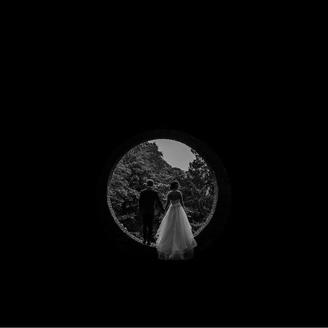 A bride and groom hold hands as they walk through a tunnel, a black and white wedding photograph.