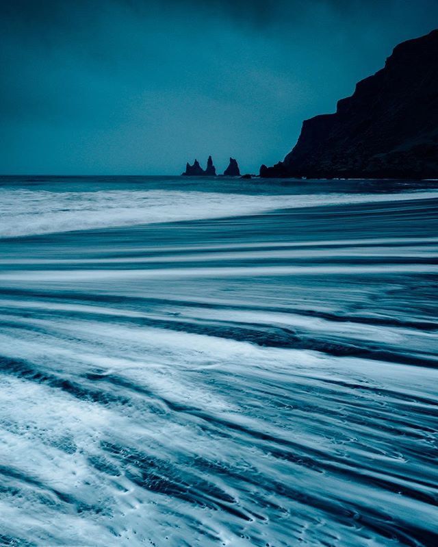 Moody seascape view of black sand beach in Iceland with dramatic ocean waves and towering cliffs on the horizon.