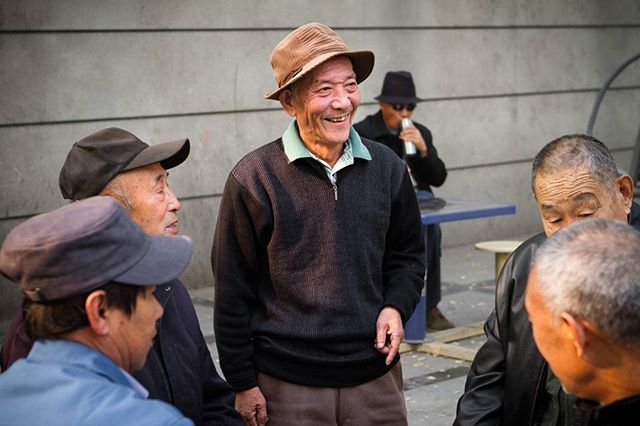 A group of elderly men gather outdoors, with one smiling broadly wearing a hat.