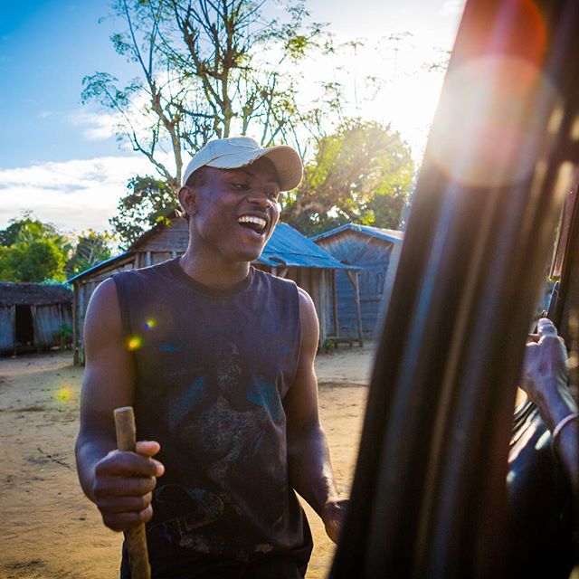 A cheerful man smiles in a rural village setting under a bright, sunny sky.