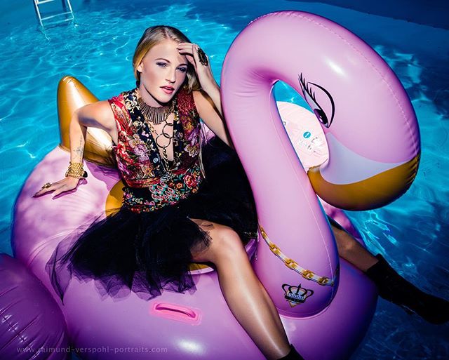 A blonde woman poses on a pink flamingo float in a pool, adorned with jewelry and a stylish outfit.