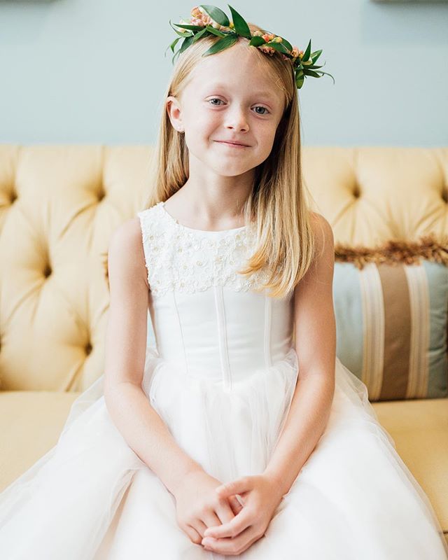 A young girl in a white dress and flower crown sits on a yellow couch, smiling gently for a portrait.