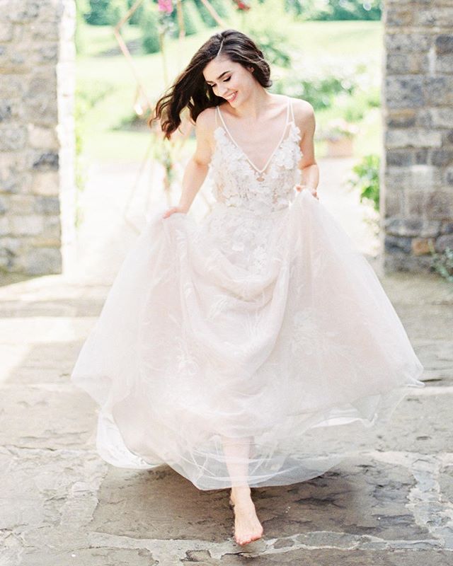A smiling bride in a flowing wedding dress walks barefoot in a garden setting.