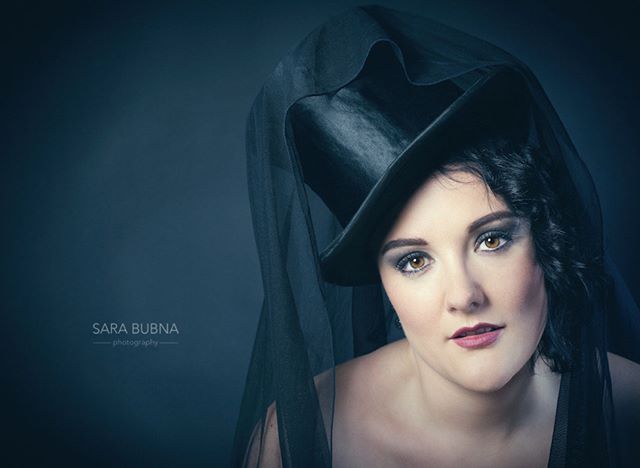 A woman in a black top hat and veil gazes intensely at the camera in a stylish portrait.