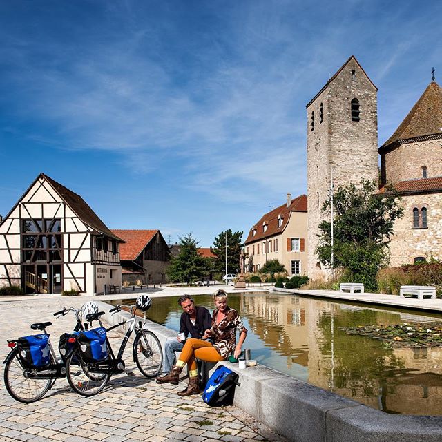 A couple relaxes by a pond in a European town square with their bicycles, enjoying a cycling tour on a sunny day.