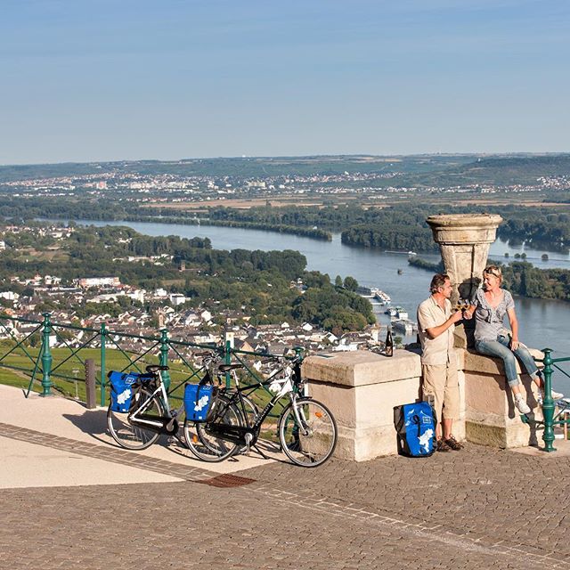 A couple enjoys wine by their bicycles, overlooking a scenic cityscape and river on a sunny day.