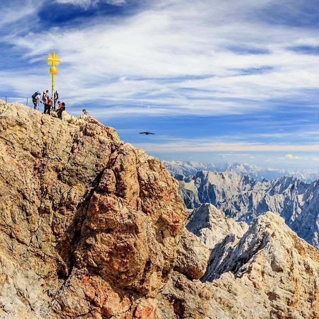 Scenic view of people at a mountain summit near a cross, with distant mountains and sky, evoking adventure and motivation.
