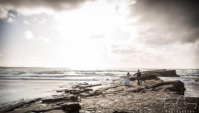A bride and groom stand on a rocky beach, the ocean behind them, captured in a romantic shot.