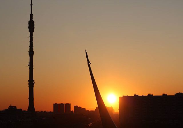 A silhouette of a city skyline with a rocket monument and TV tower against a warm sunset.