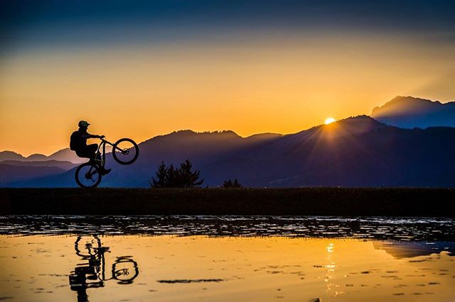Silhouette of a mountain biker doing a wheelie at sunset with mountains and a water reflection.