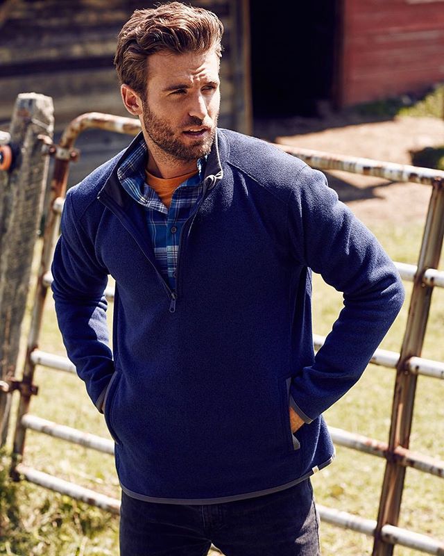 A man in a blue fleece and flannel shirt poses outdoors in a rural setting.