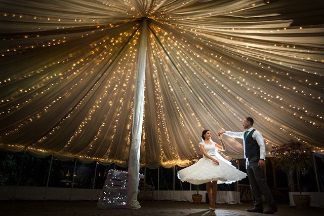 A bride and groom share a dance at their wedding reception inside a tent lit with warm fairy lights.