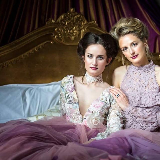 Two women pose in elegant vintage dresses on a golden bed with purple drapery. Editorial fashion.